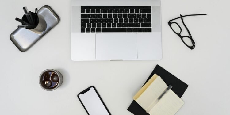 Top view of modern laptop and smartphone with empty screens placed near opened notebook and eyeglasses on light table